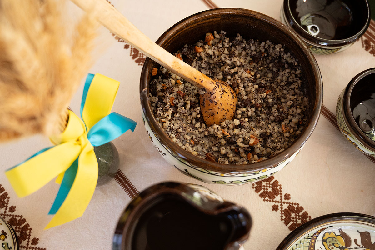 From above, a ceramic bowl of a wheat berry porridge dotted with almonds and poppy seeds, a ceramic carafe if dark liquid, empty bowls and dishes, and a bouquet of dried wheat stalks with with yellow and blue ribbons - the colors of the Ukrainian flag.
