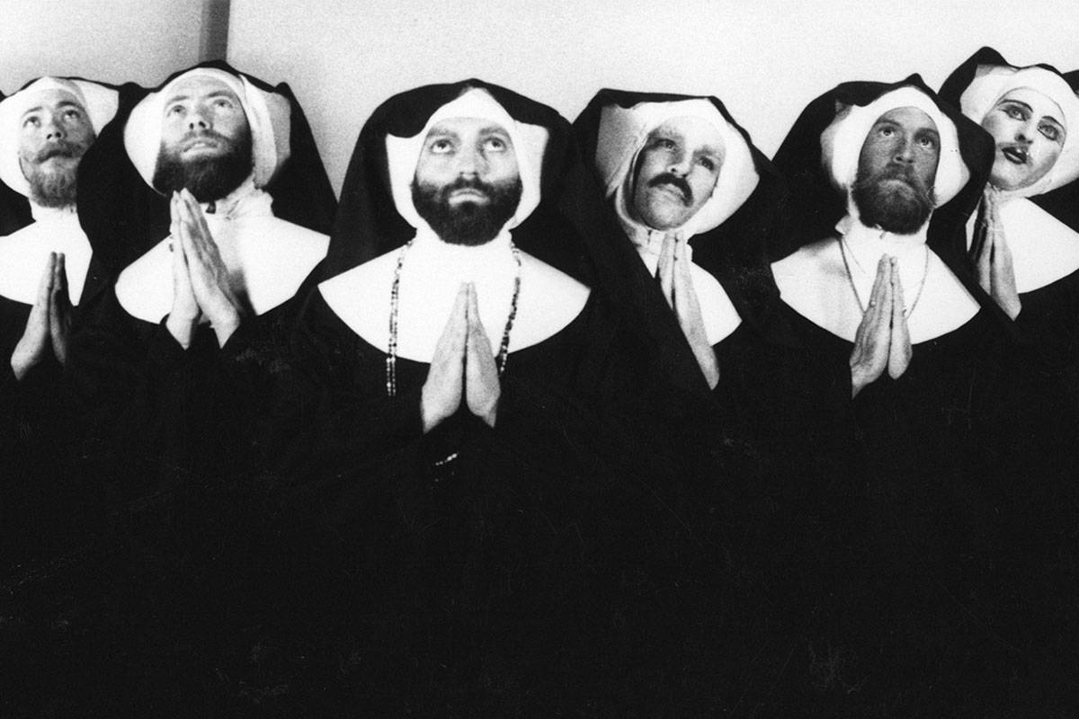 Six people dressed in black and white nuns' habits look upward and bring hands together in prayer. Black-and-white photo.