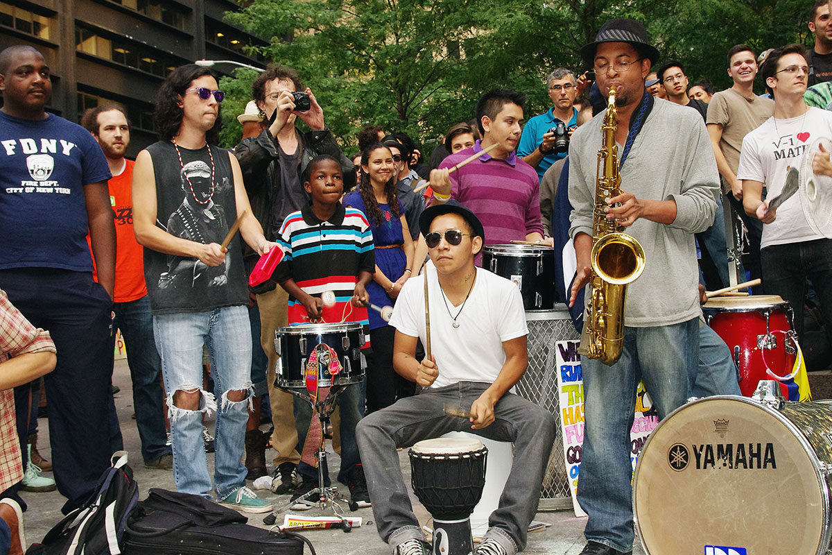 Crowd of people playing music outside, with others around them observing. Most people play some kind of percussion; one person plays saxophone.