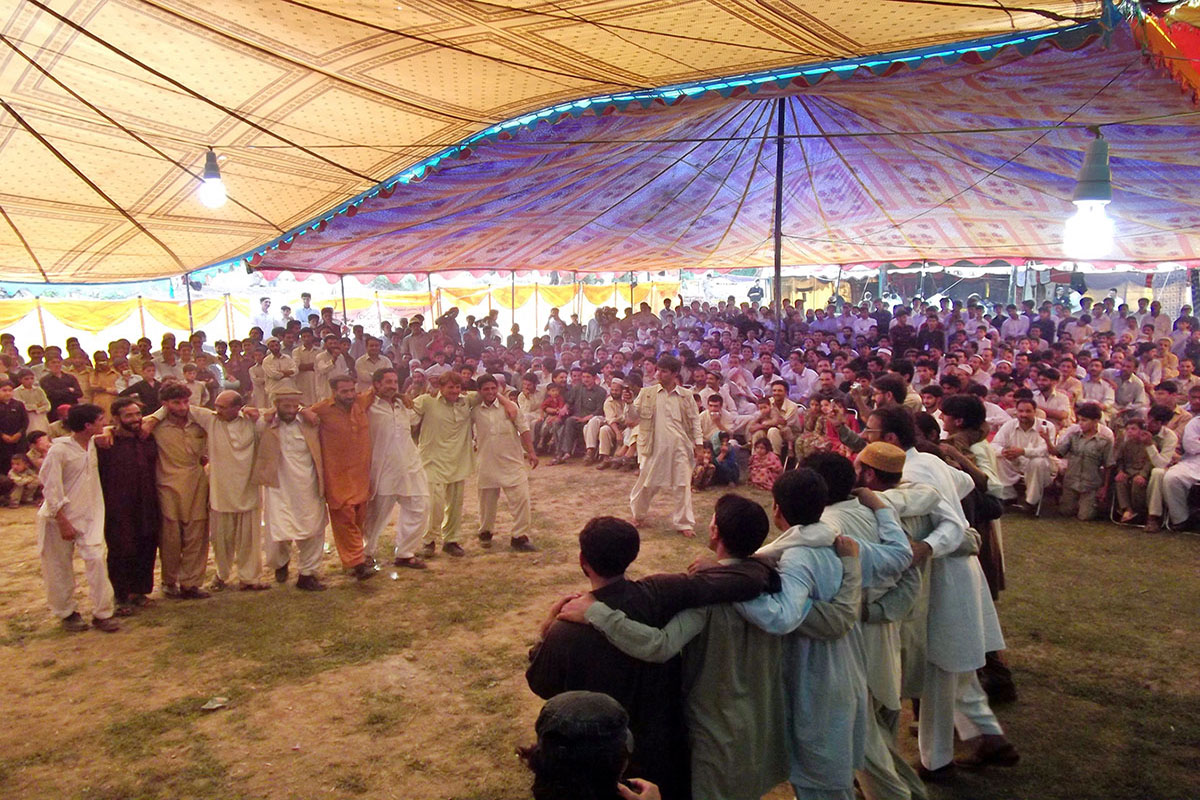 Two groups of men shoulder-to-shoulder face each other in traditional dance for a crowd under a large cloth tent. 