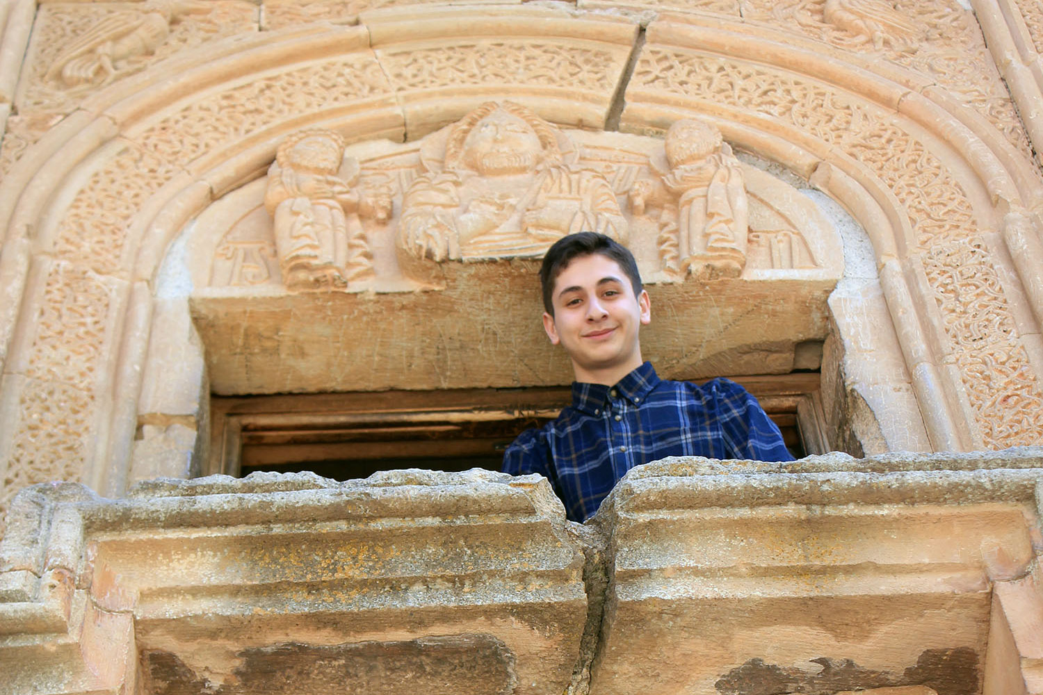 A teenage boy looks down at the camera through an open window of a beige, ornately carved stone monastery.
