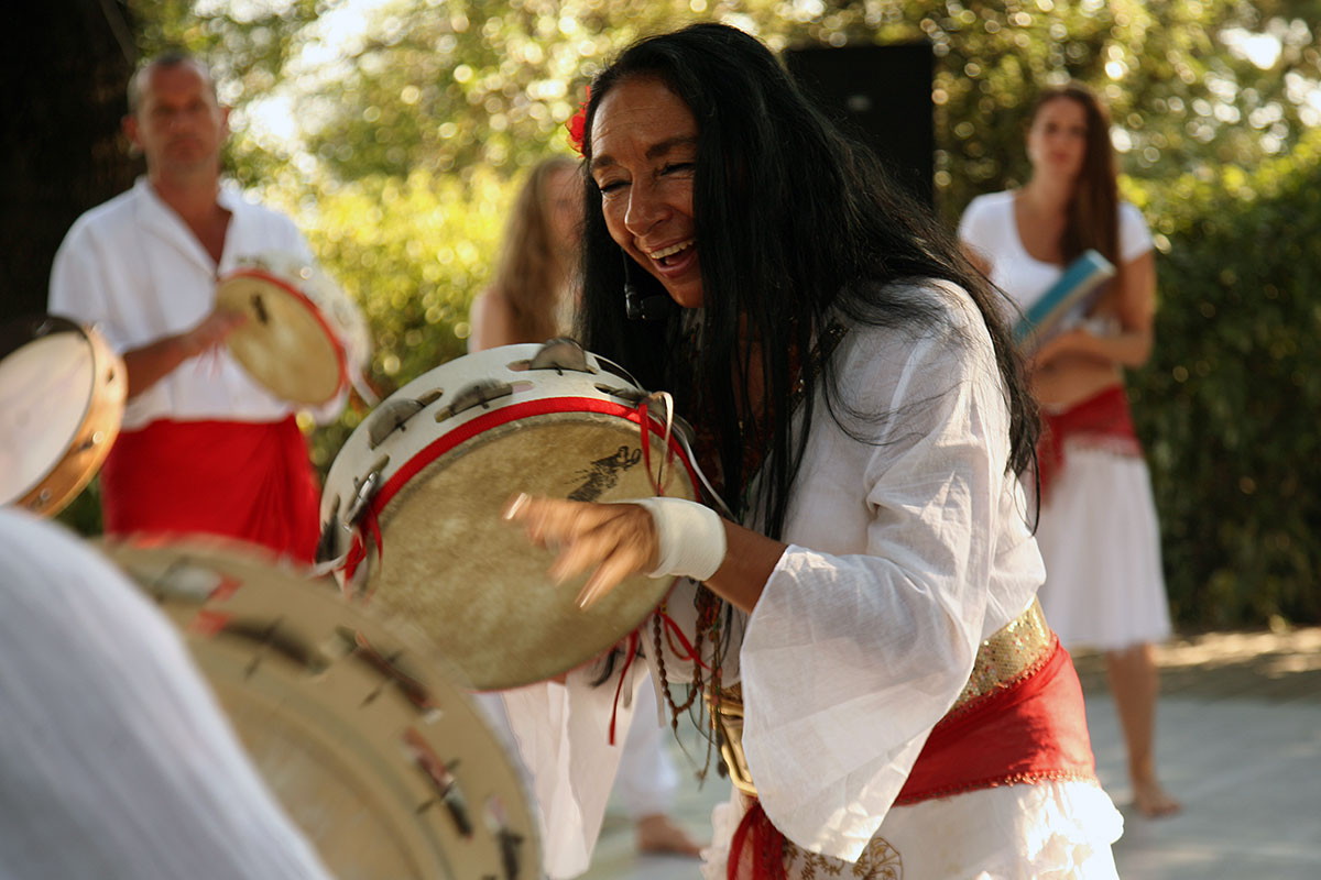 A woman in flowy white blouse and red sash plays a wooden frame drum with her hands, demonstrating for others in the same outfit around her.