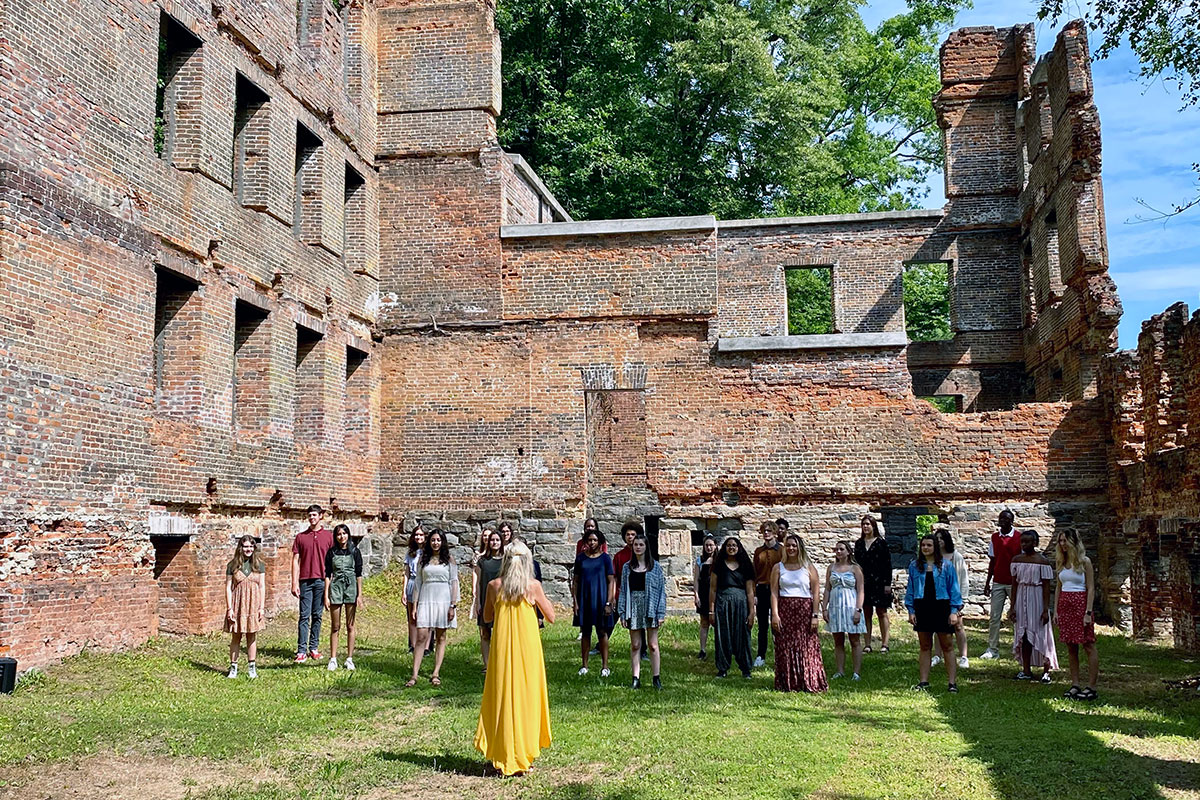 Rows of teens stand in formation, socially distanced, with a woman in a bright yellow dress standing in front, conducting them. They are outdoors, surrounded by the remnants of a dilapidated, roofless brick building. 