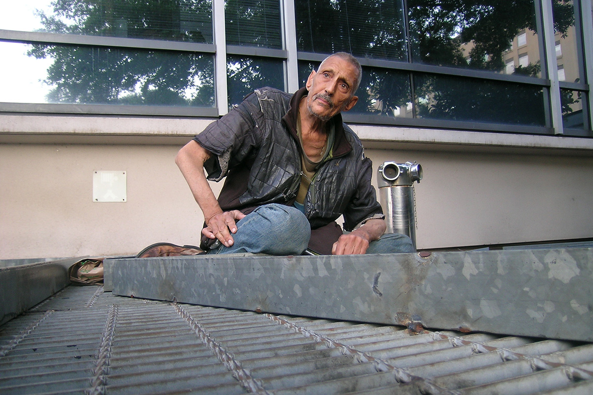 A man in tattered clothes looks pensive, sitting atop a heating grate at the edge of an office building.