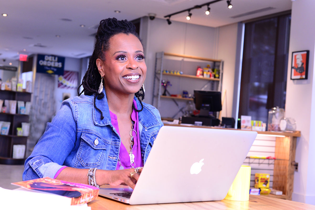 A Black woman sits working on a laptop at a café/deli, smiling into the distance. 