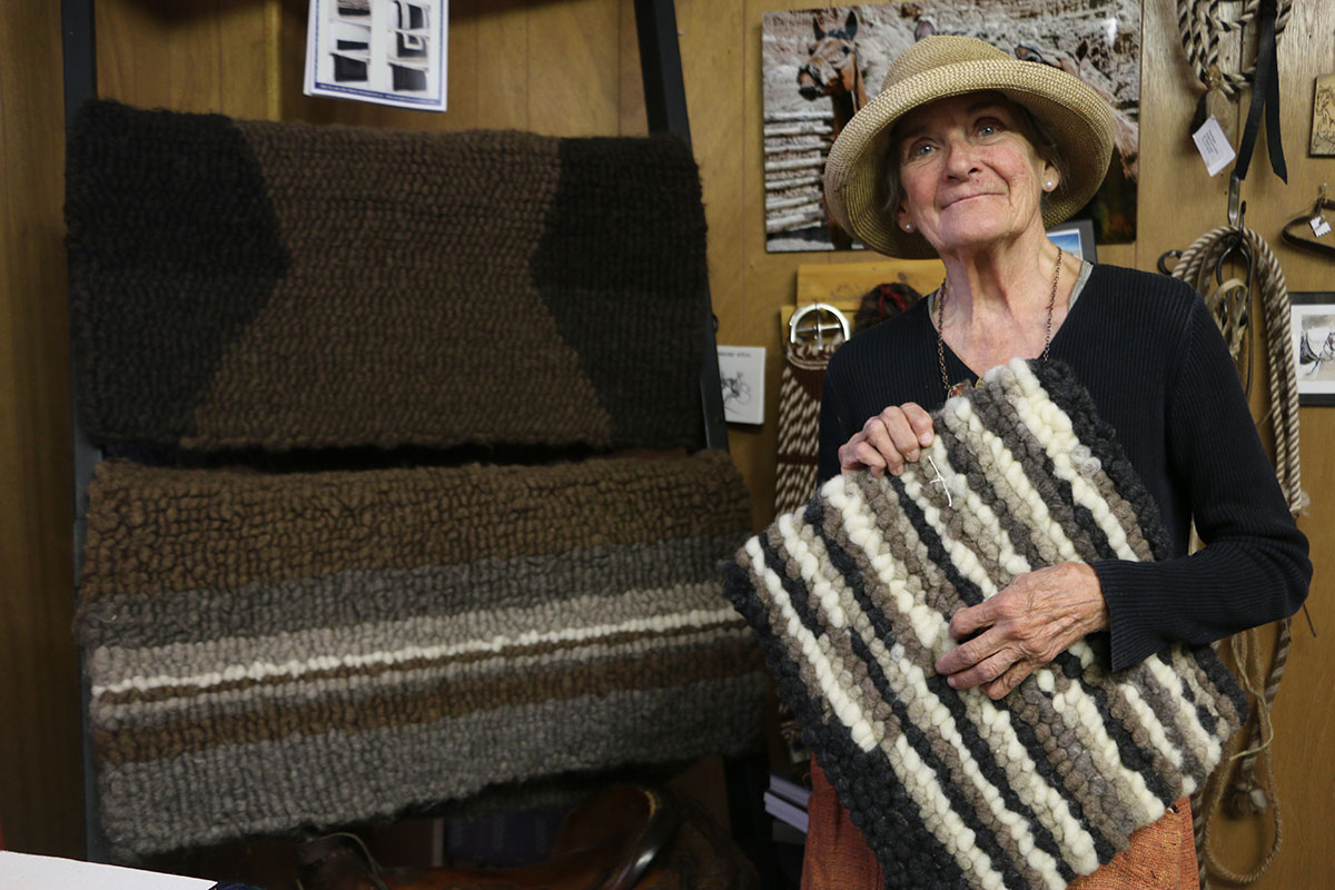W woman holds up a saddle pad: a rectangular textile in stripes of white and brown tufted wool. Other saddle pads hang on a wall rack next to her.