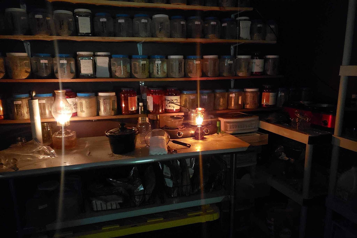 Wooden shelves and counter filled with jars are lit by two oil lamps.