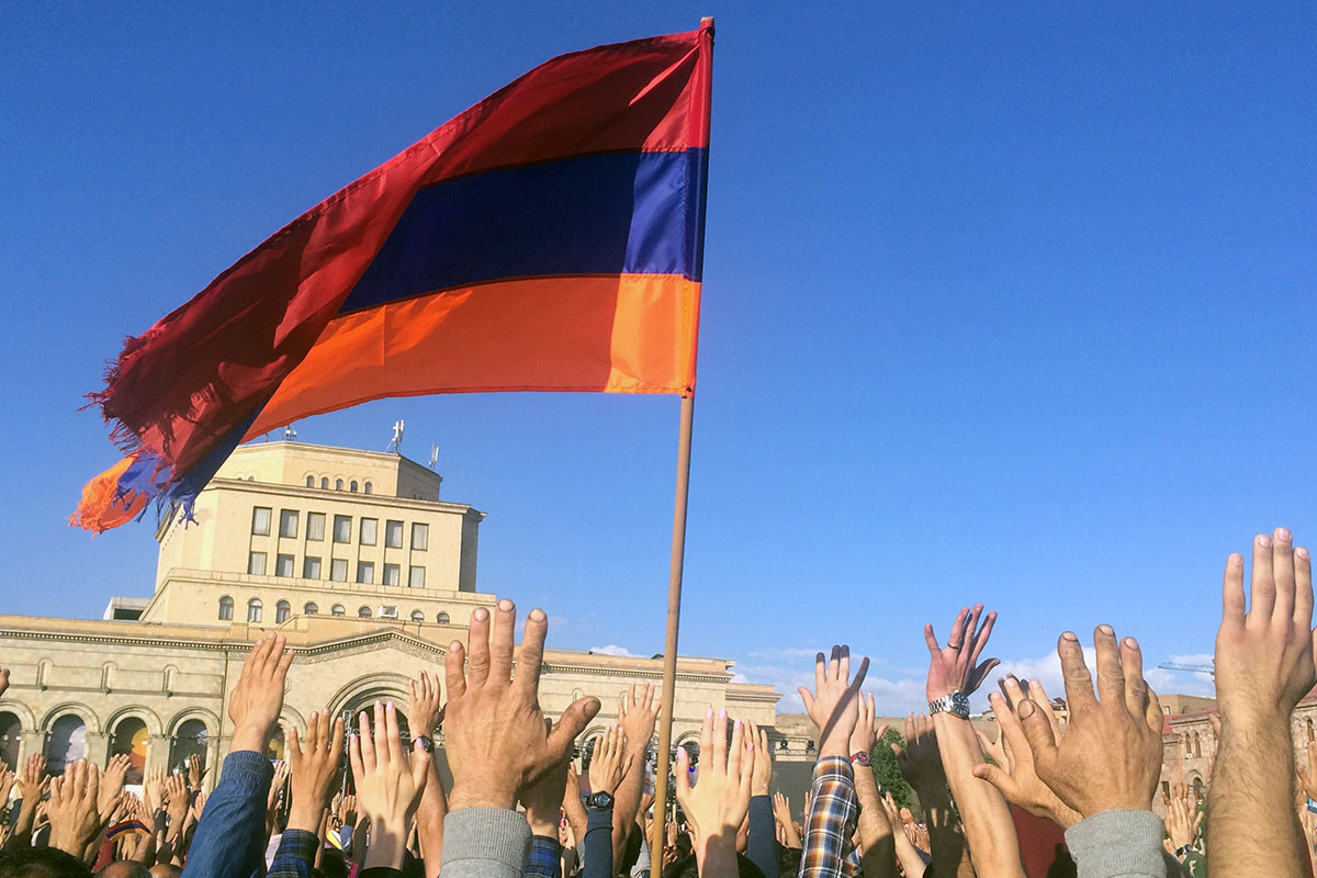 The Armenian flag (with three stripes of red, blue, and orange) is raised along with hundreds of hands under a blue sky, with a government building in the background.