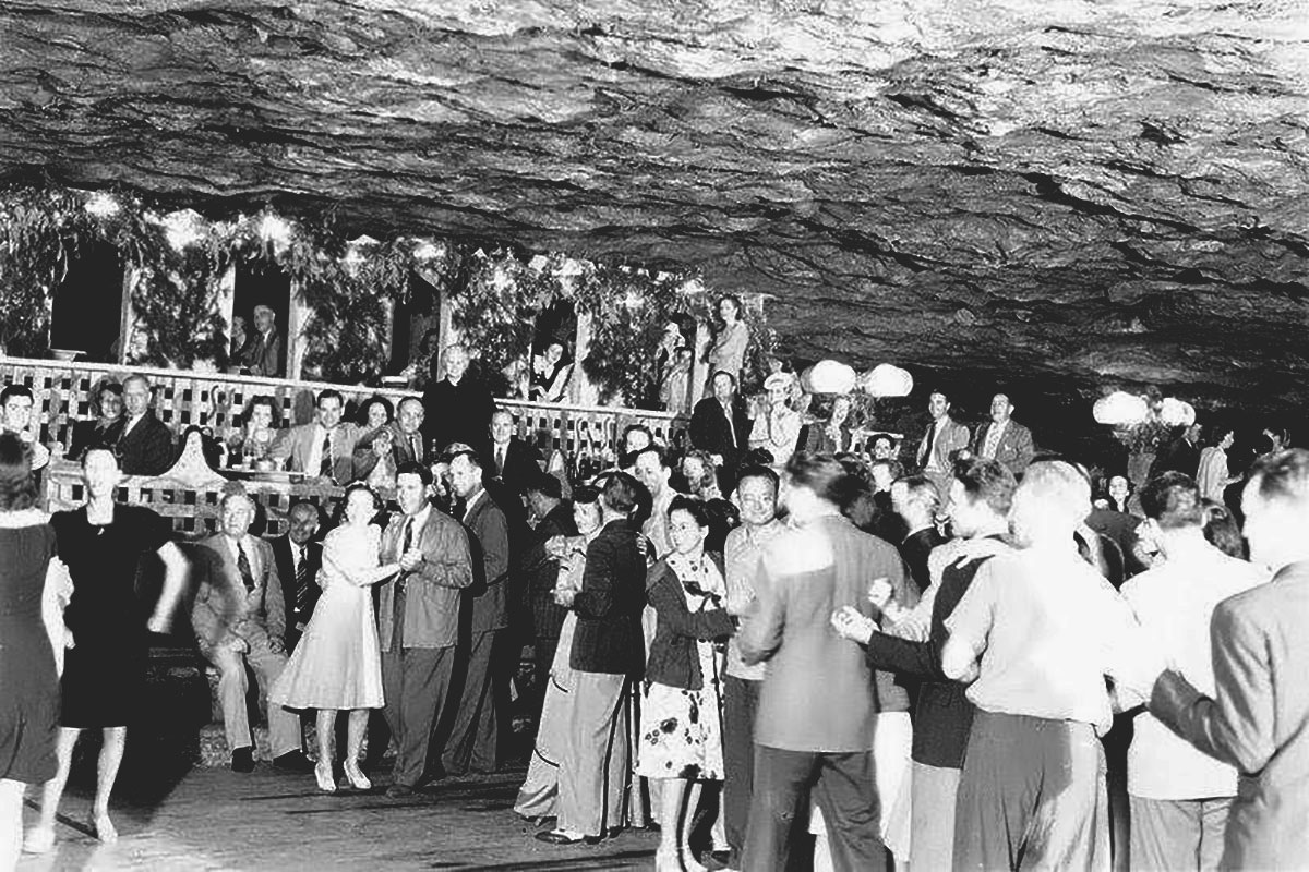 Dozens of people dressed in evening wear dance and enjoy an event inside a cave, under a rocky ceiling. Black-and-white photo.