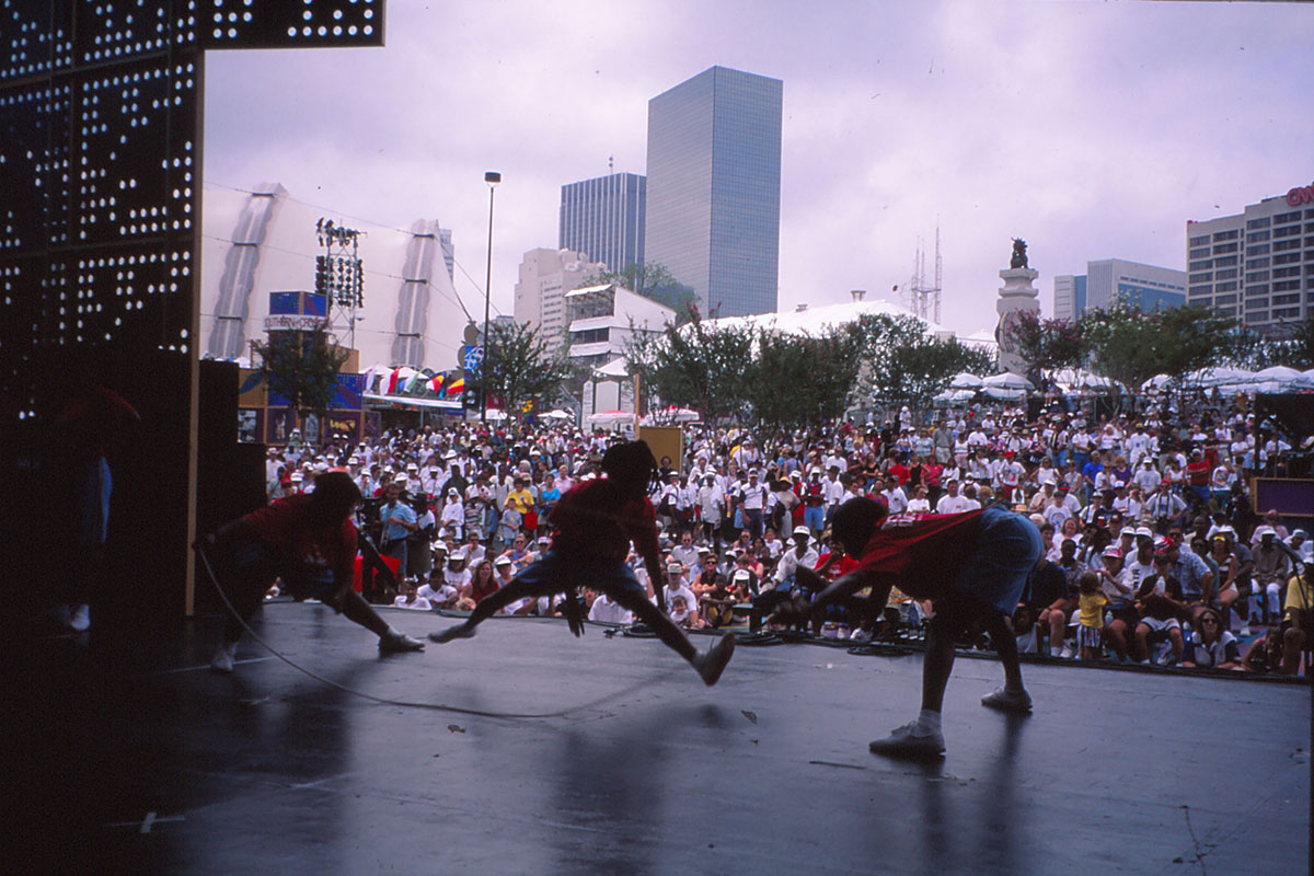 From the back of an outdoor stage, three young people do double-dutch jump rope for a huge crowd of people. Skyscrapers in the background.