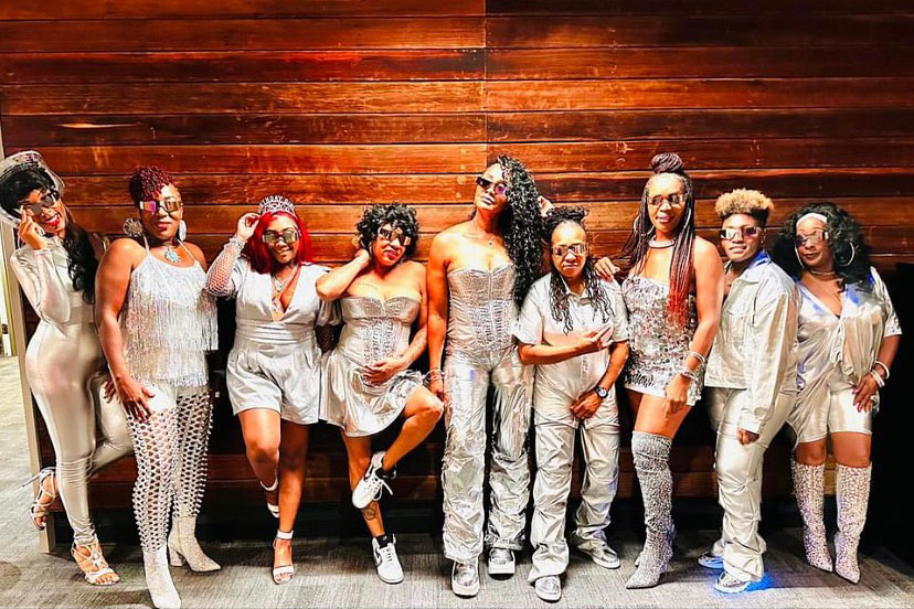 Nine Black women dressed in varying silver outfits and sunglasses pose in front of a wood-paneled wall.