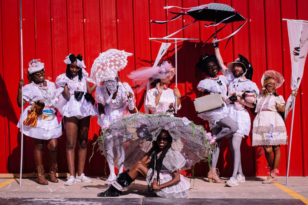 Eight people, both women and men, pose against a red wall, each dressed in a frilly pale pink dress with bonnets or large bows on their heads. Some carry decorative parasols. Some carry small dolls. 
