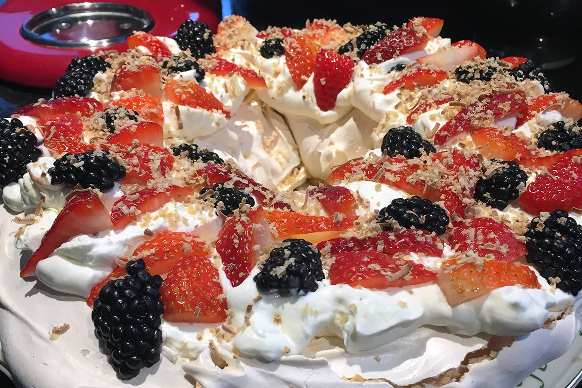 Close-up on a round pavlova: white meringue topped with soft whipped cream, sliced red strawberries and dark blackberries and small chocolate shavings.