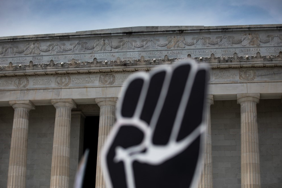 A black foam fist raised in the foreground, with the Lincoln Memorial and clear blue sky in the background.