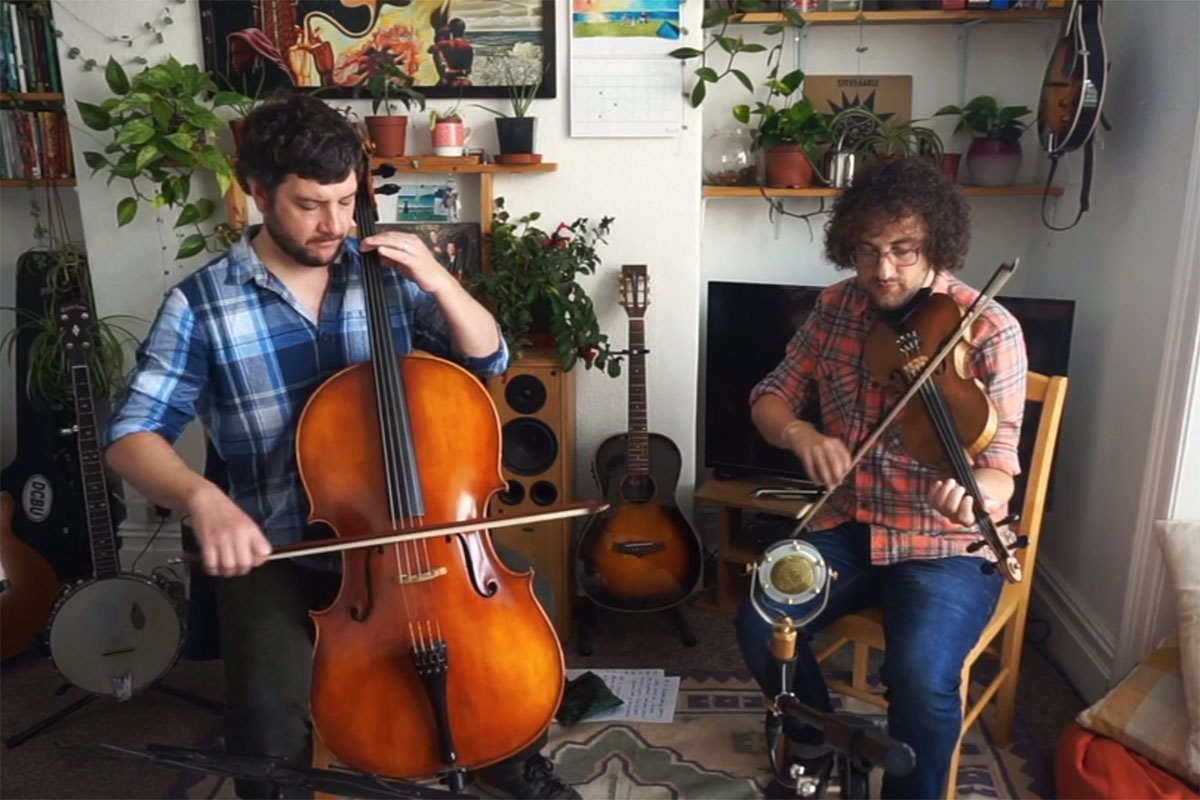 Two musicians playing in a living room setting, one on cello and the other fiddle. Other stringed instruments are on the floor and hanging on the wall behind them.