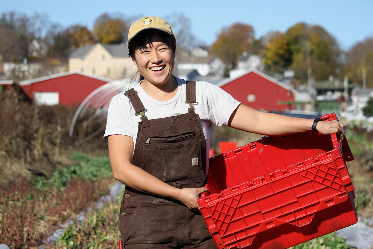 A young Asian woman in white T-shirt and  brown overalls holds a red plastic container, smiling at the camera, with a farm behind her.