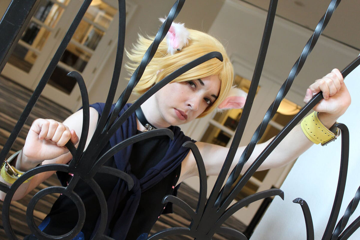 A person in a costume of black tank top, short yellow wig with furry white ears, and yellow leather bracelets, poses through a black wrought-iron railing.