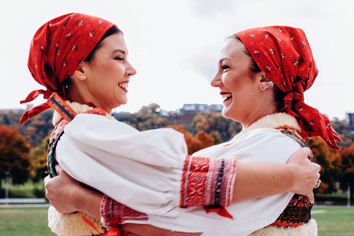 Two women in matching white blouses with red embroidered cuffs and red headscarves embrace each other in dance, smiling. Trees in the background are beginning to turn autumn colors.