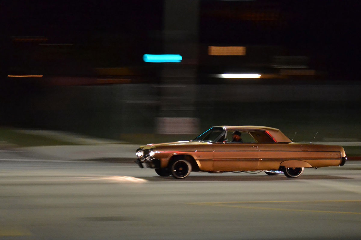 At night, a person drives a classic car down a street, street lights blurred in the background.