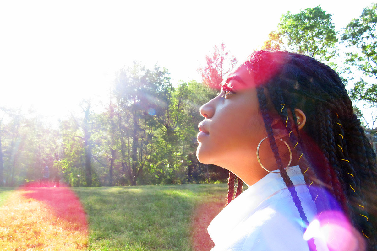 A Black Latinx woman with long braids and gold hood earrings standing in a park, with red and purple spots of lens flares.