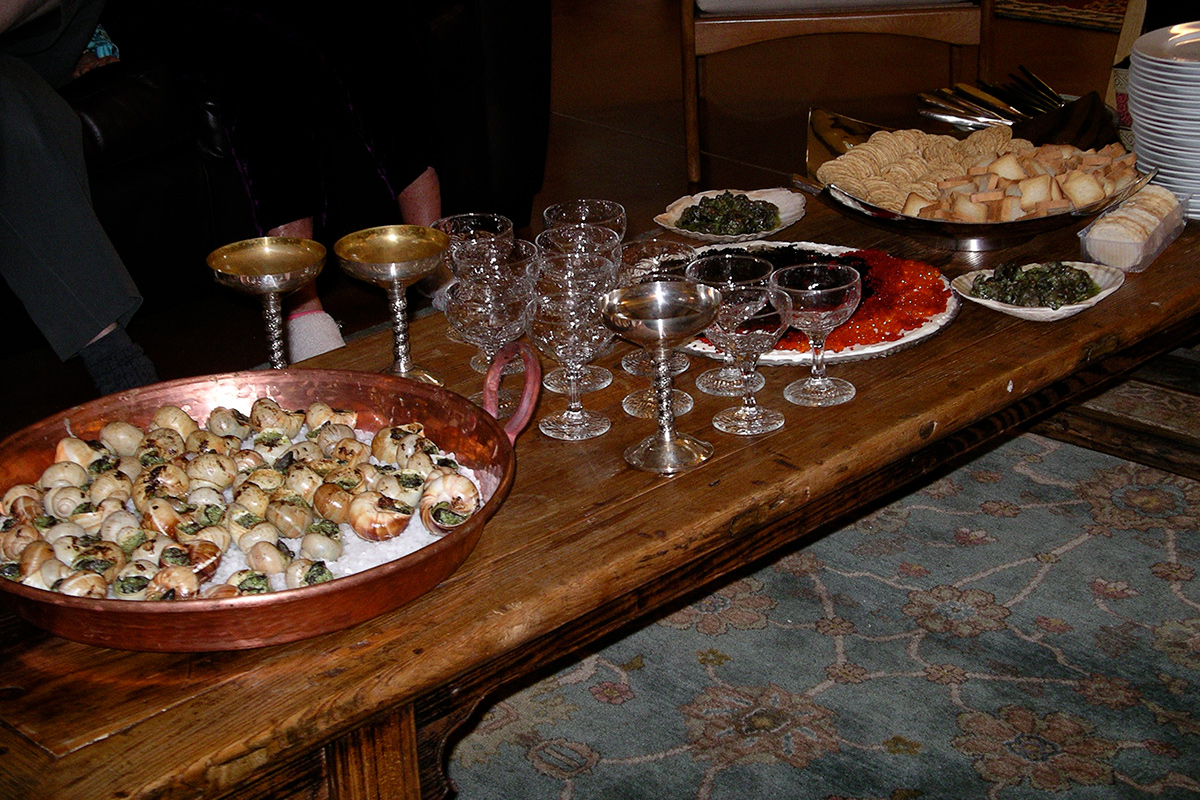 A coffee table set with a copper pan full of escargots on rock salt, wide champagne glasses, a plate of red and black caviar, and a bowl of crackers.