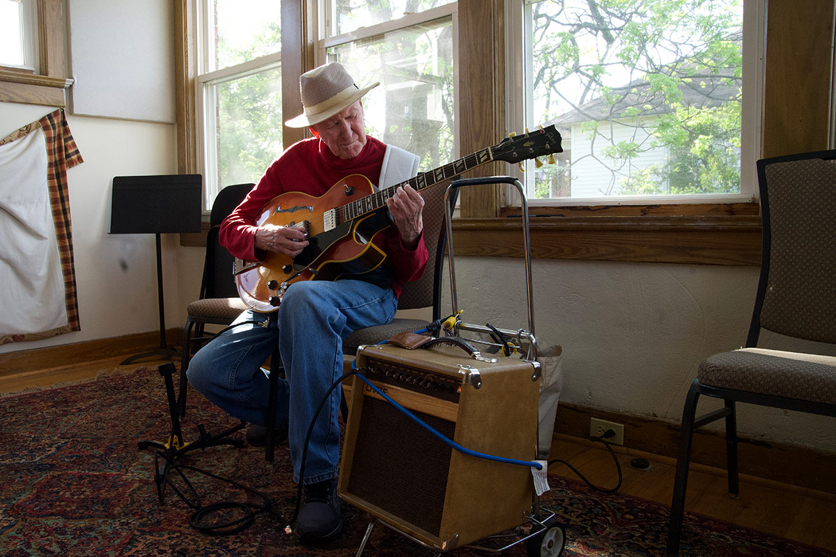 A man wearing a tan fedora, red long-sleeve-shirt, and blue jeans plays an electric acoustic guitar in a practice room, next to windows with sunlight streaming in.