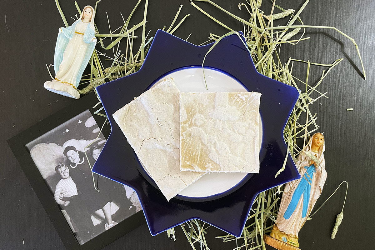 Two pale, rectangular wafers on a decorative plate, set besides dry hay, two Mary figurines, and a framed family photograph.