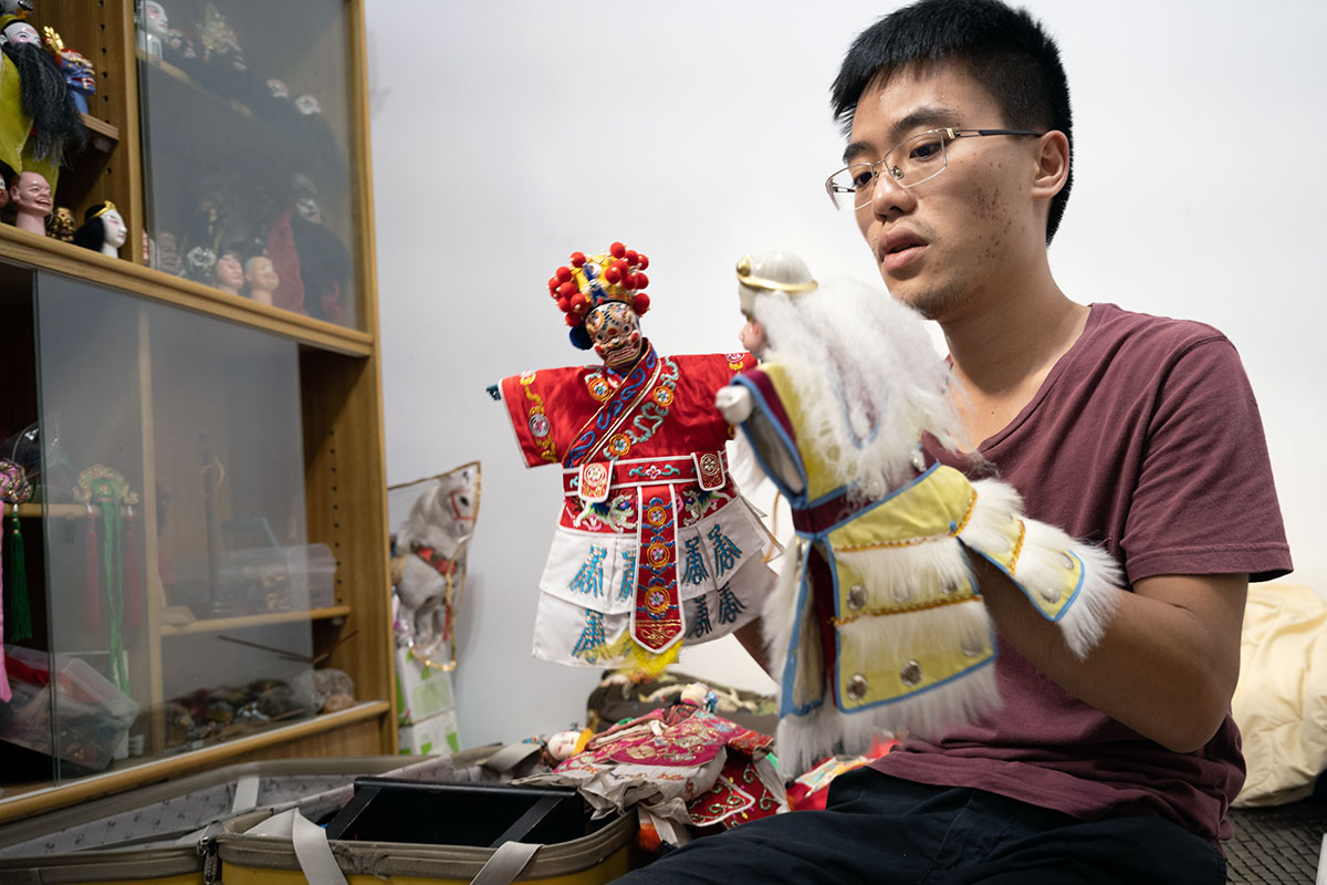 A young man, seated, holds glove puppets on hand, both intricately costumed in Chinese dress. A cabinet behind him holds more puppets and puppet pieces.