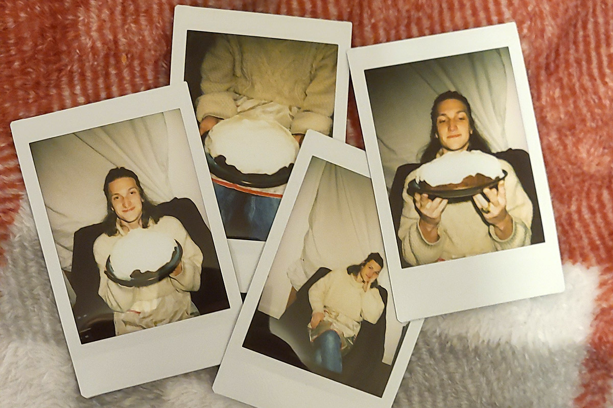 Four Polaroid photos of a person posing with a brown cake with white frosting. The photos are laid out on a white and red plaid blanket.