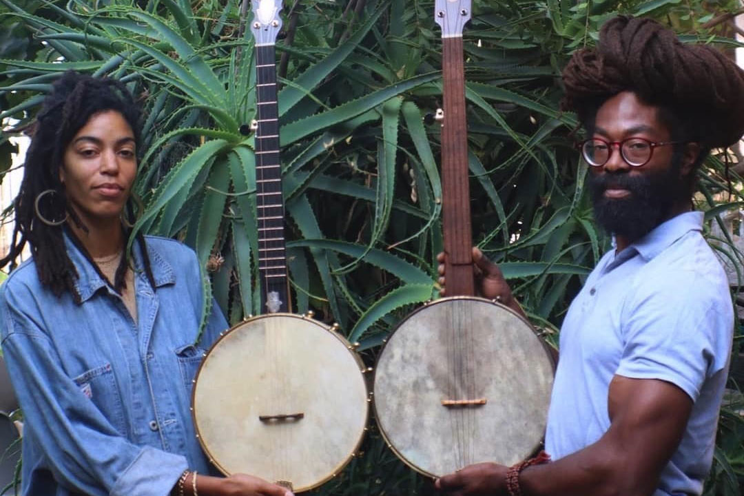 A woman and a man, both Black, hold up banjos in front of a large succulent.