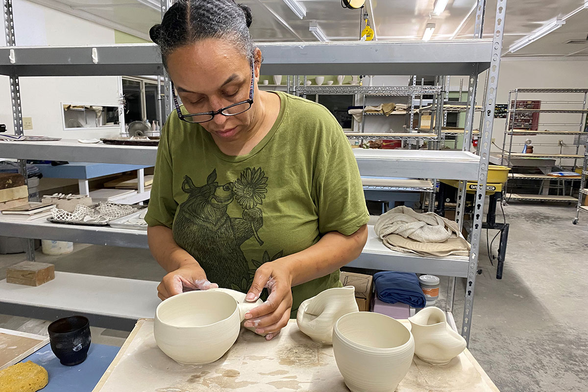 A Black woman forms a clay bowl with her hands. Other dishes in progress sit on the table in the foreground. 