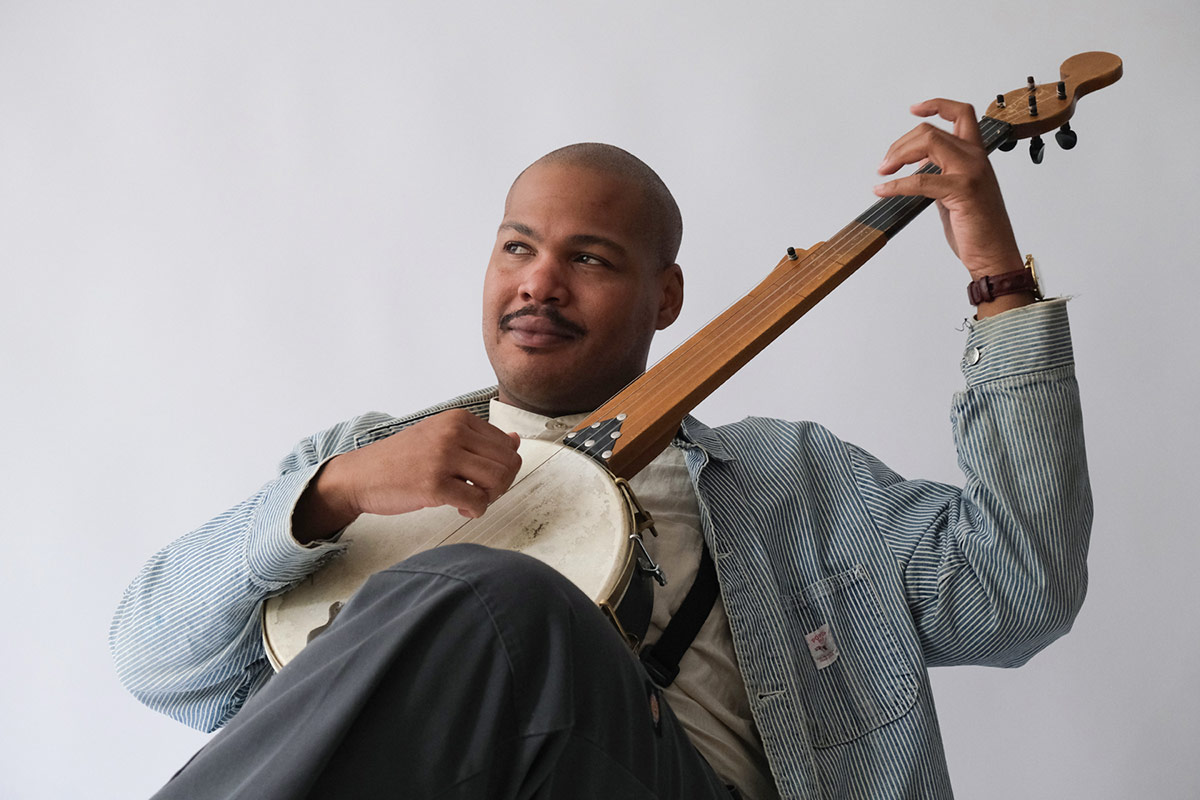 A Black man reclines, playing a banjo and looking into the distance.