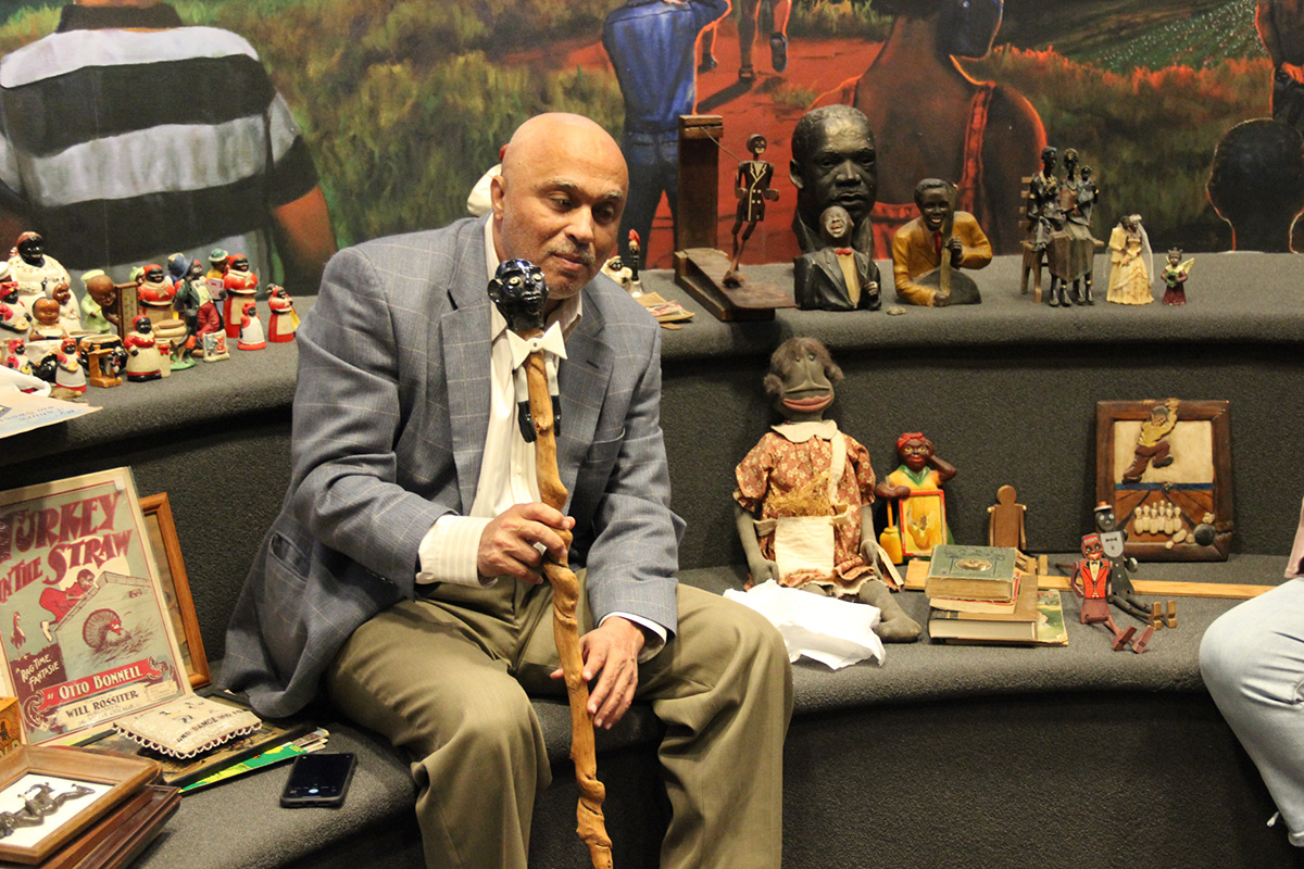 A man sits surrounded by racist objects, using Black caricatures on dolls, a cane, posters, and more.