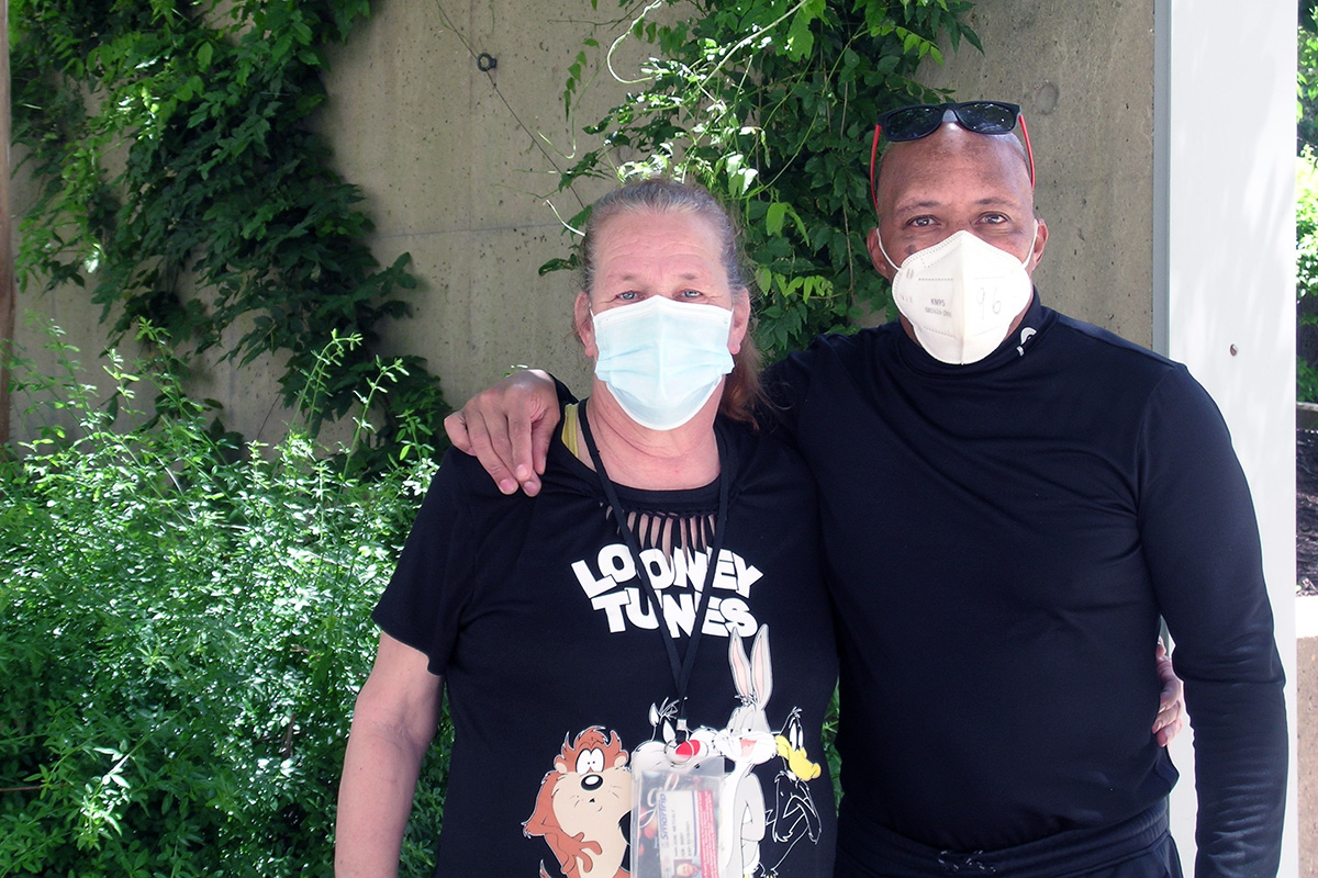 A husband poses with his arm around his wife, both wearing medical masks. He is black, and she is white. She wears a Looney Tunes shirt showing the Tasmanian Devil, Sylvester the Cat, Bugs Bunny, and Daffy Duck.