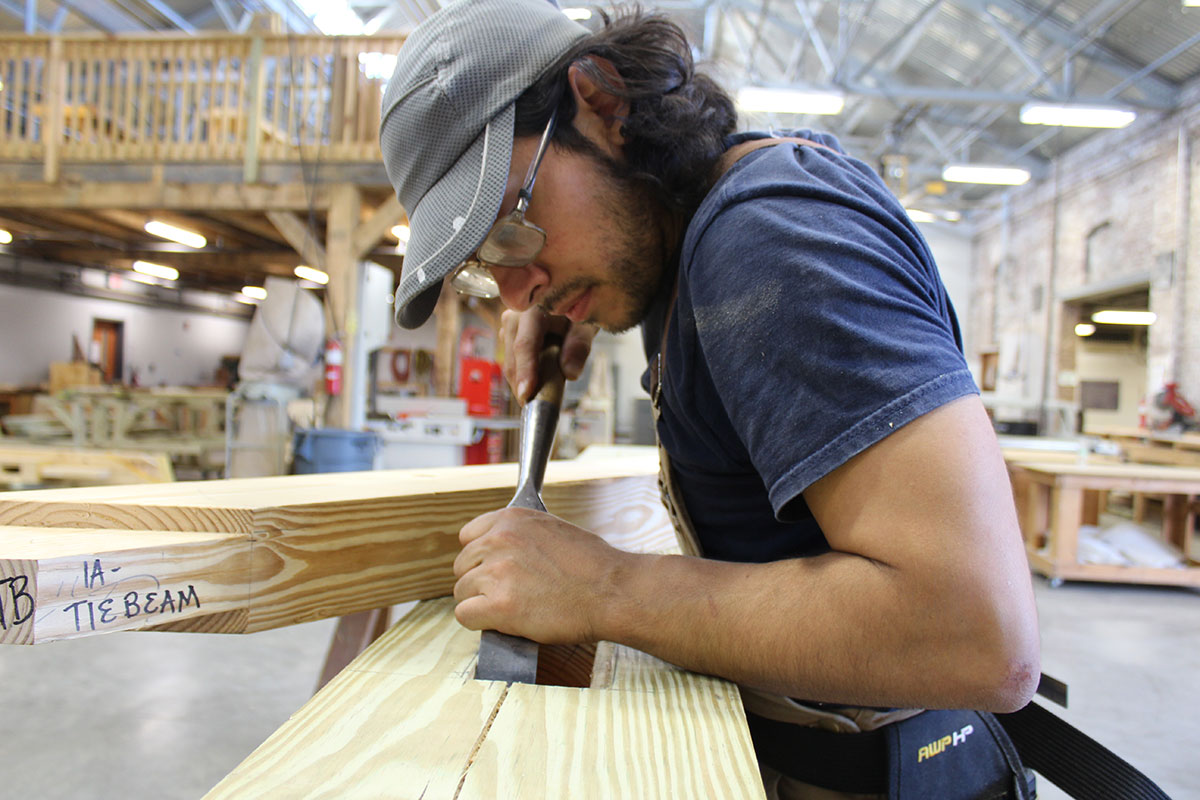 A young man fine tunes a cut in a piece of lumber using a hand tool that looks like a wide chisel.