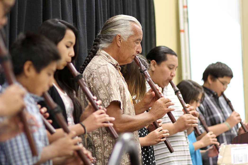 A row of people lines up playing flutes. Most are young students, and in the middle is an older man with a long gray braid.