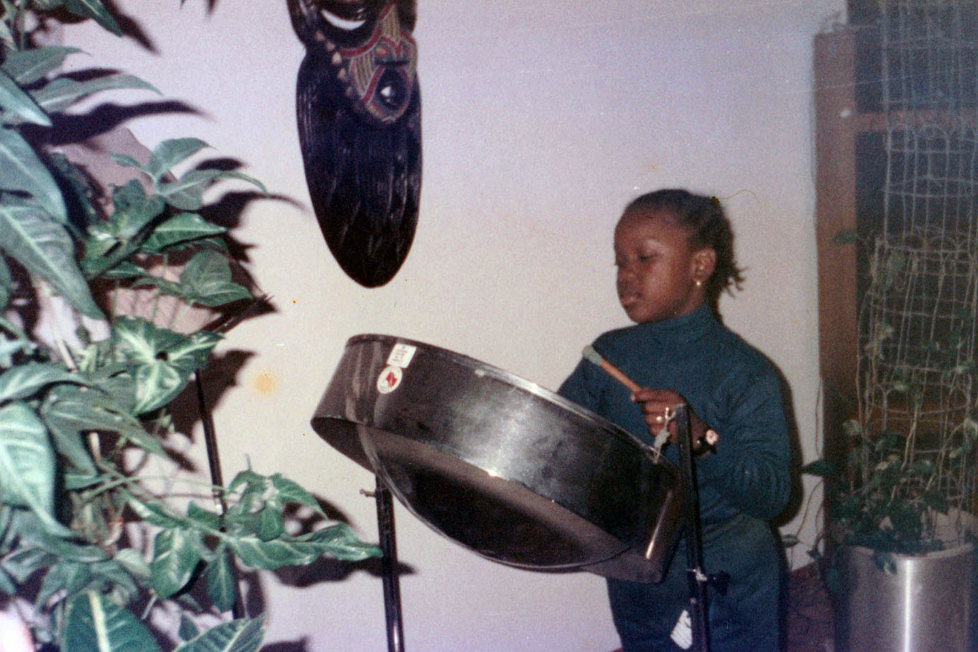 A young girl plays a steelpan drum, houseplants in the foreground and background, and a wooden mask on the wall.