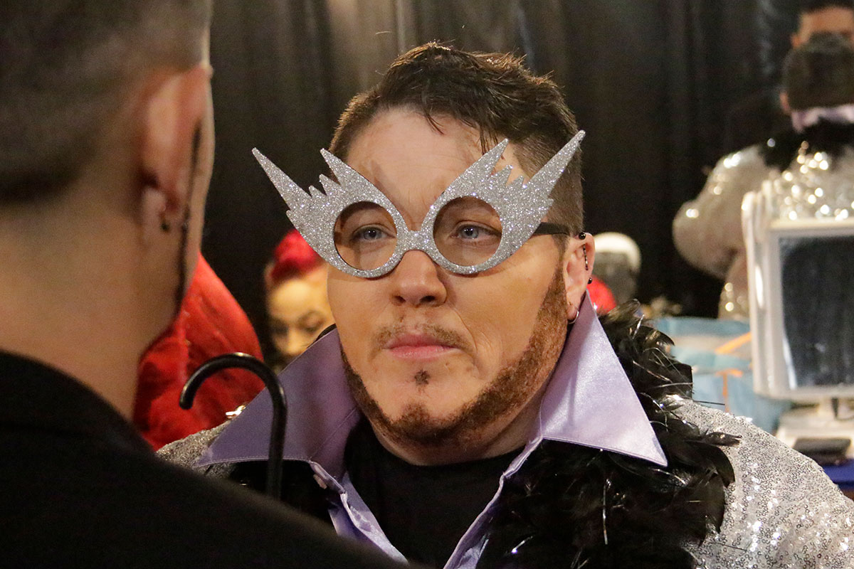 A person with glittery silver coat with oversized lavender collar, black feather  boa, prop glasses - no lenses but silver frames that resemble comets with jagged trails behind them - and a thin faux beard faces another person in a mirrored dressing room.