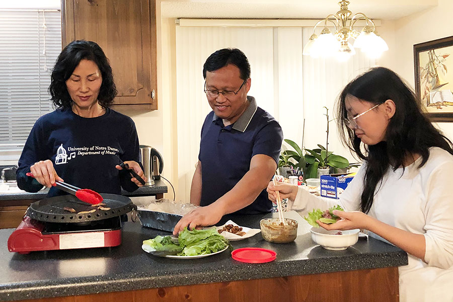 Three people stand around a kitchen island counter. The mom is using tongs to cook something on a hotplate. The dad is reaching for a plate of greens. The daughter is using chopsticks to fill a lettuce leaf with a brown filling.