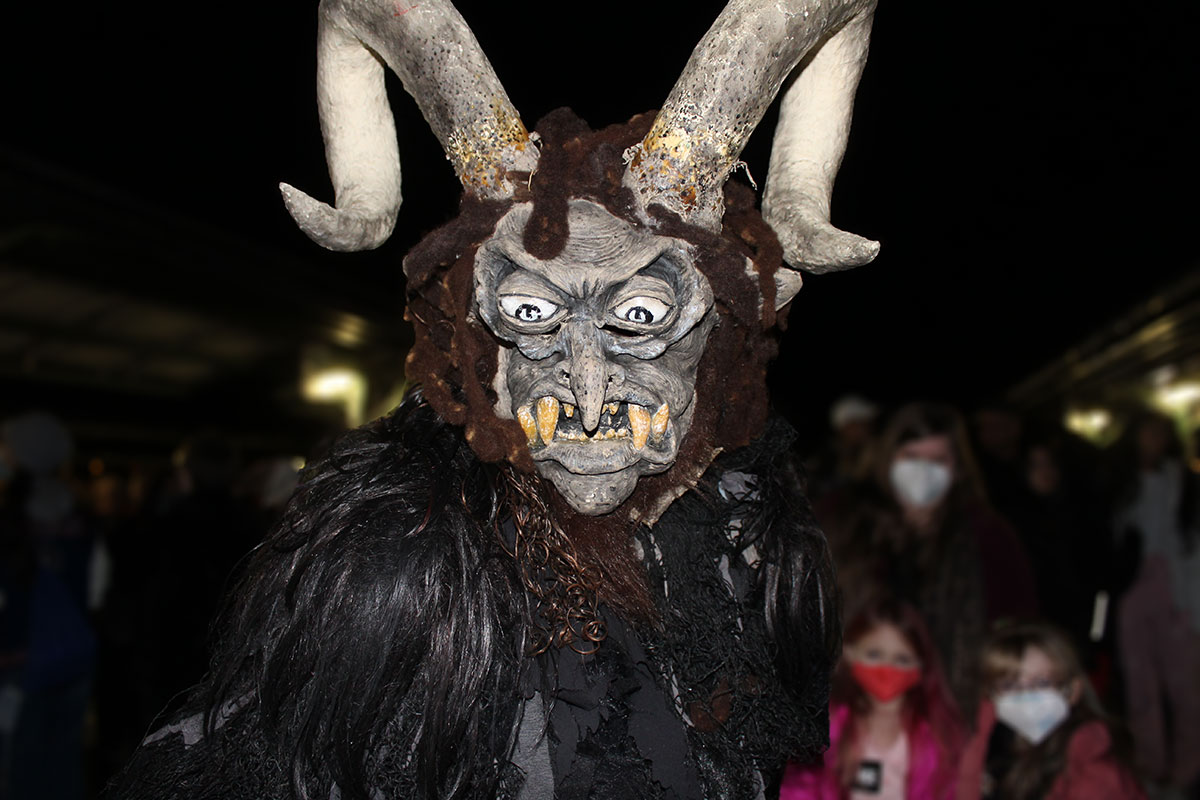 A person in a Krampus costume with bulging eyes, curved horns, and protruding teeth. Two young kids in face masks and an adult watch the Krampus.