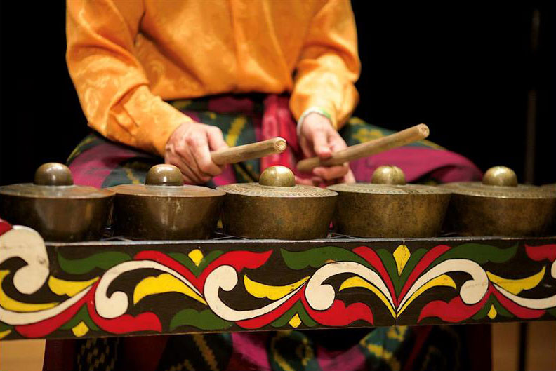 A person (face unseen) holding two wooden beaters plays a musical instrument of several gongs facing upward, sitting in a wooden frame that is painted in black, green, yellow, red, and white.