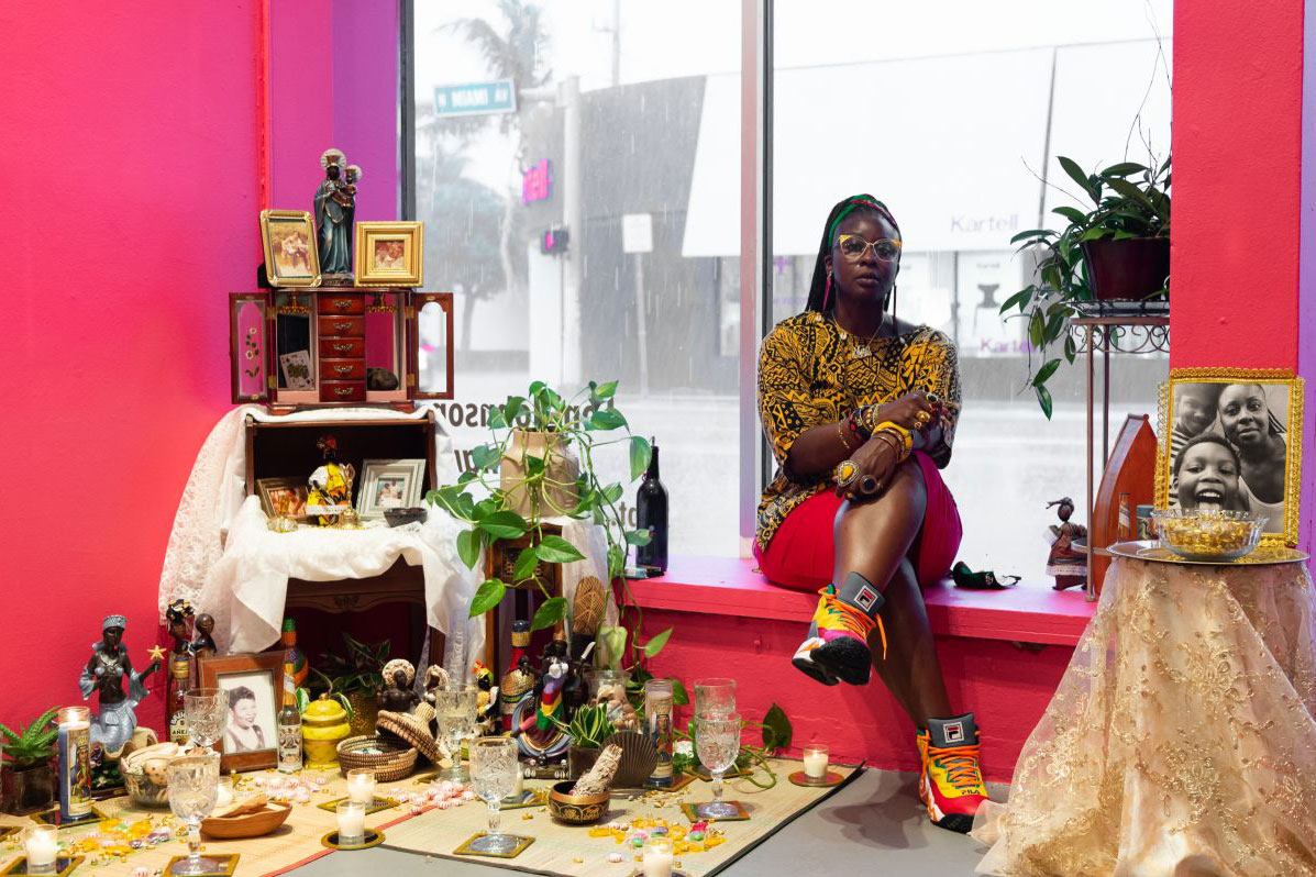 A Black woman with yellow and black printed blouse and fat sneakers sits in a window sill among pieces of an art installation, an altar with framed photos, a Black Madonna figurine, green potted plants, crystal glasses, and more.
