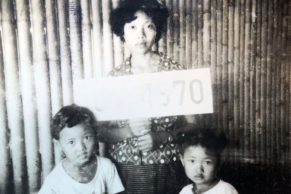 A mother and two young children pose in front of a bamboo wall. The mother folds a white sign with a number, washed out by light. Black-and-white photo.