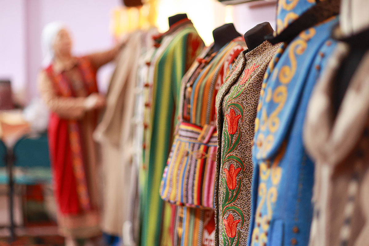 In the foreground, a lineup of mannequins each with colorful embroidered garments. At the end of the line, out of focus, a woman dresses a mannequin.