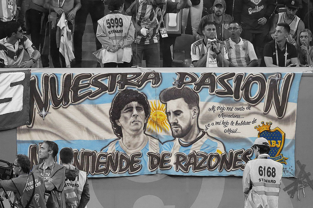 Banner in a soccer stadium with portraits of Maradona and Messi over the colors of the Argentina flag. The banner is in color, while the seated fans behind and film crew in front are in black and white.