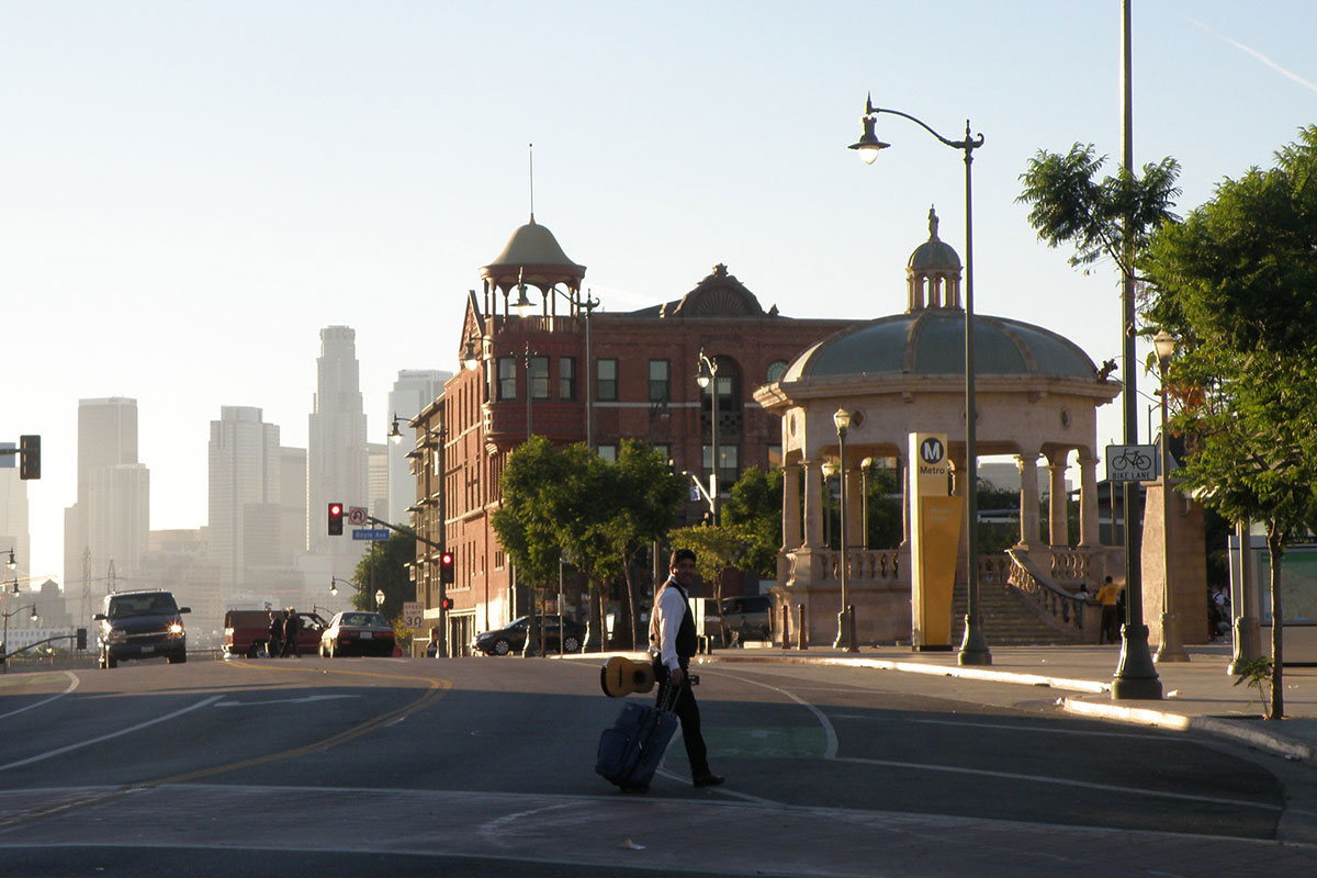 A man holding a small guitar and a suitcase crosses a street toward a large gazebo. The towering Downtown Los Angeles skyline looms in the distance.