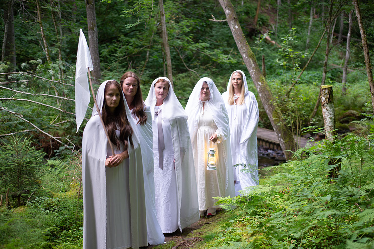 Five adults dressed in white dresses, robes, and veils pose along a forest path. One carries a lit lantern, and another carries a white flag.