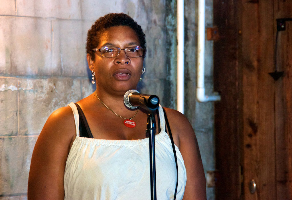 A Black woman with short natural hair, white tank top, and red dogtag necklace speaks into a microphone.