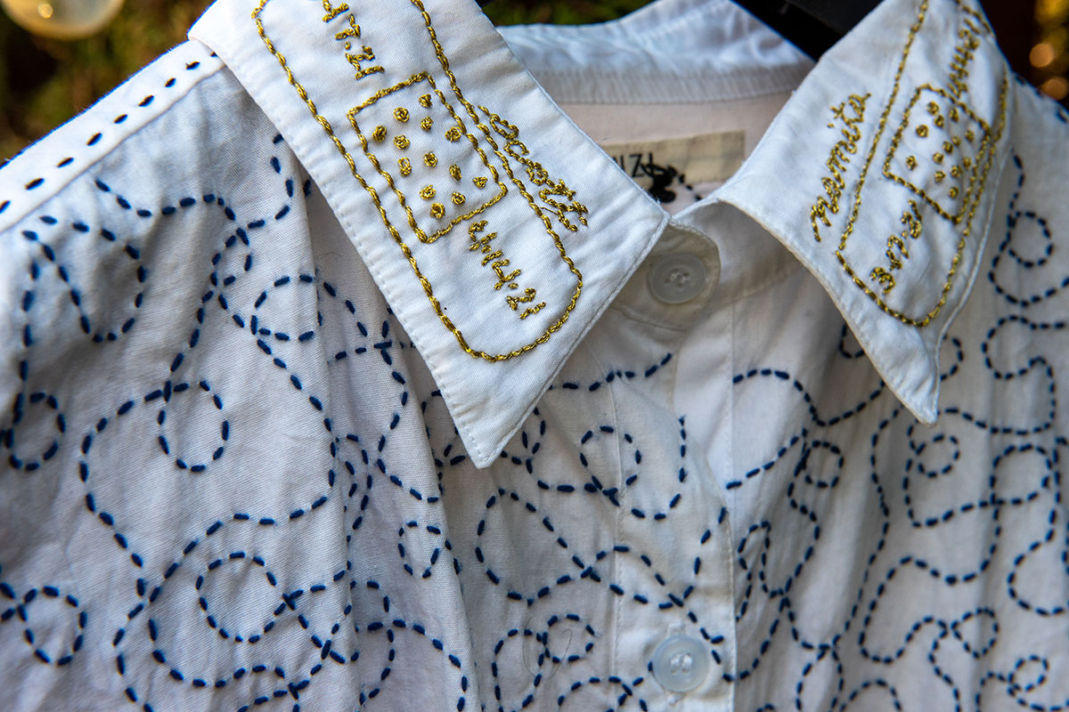 White button-up shirt embroidered across the body with a continuous blue squiggly line. On the collar are two embroidered Band-Aids with the names Scott and Namita and two sets of dates. 