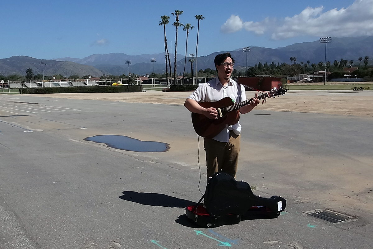 A man sings and plays guitar on a wide blacktop, with a few palm trees in the near distances and mountains afar.